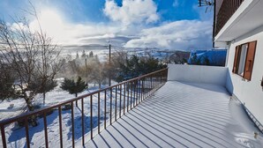 A snow-covered deck with a metal railing, a view of a snowy landscape, and a building with a white facade and brown window frames.