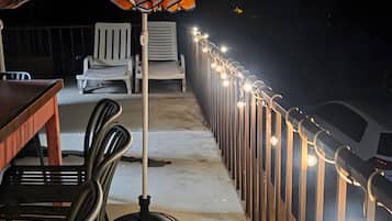 Uma área de varanda ao ar livre com mesa e cadeiras, um guarda-sol listrado e um corrimão com luzes.