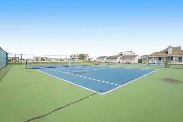 An outdoor tennis court with a net, surrounded by a fence, and residential buildings in the background. - Bethany Bay 2nd floor condo w/shared pool, tennis court, on-site golf, balcony (Ocean View)