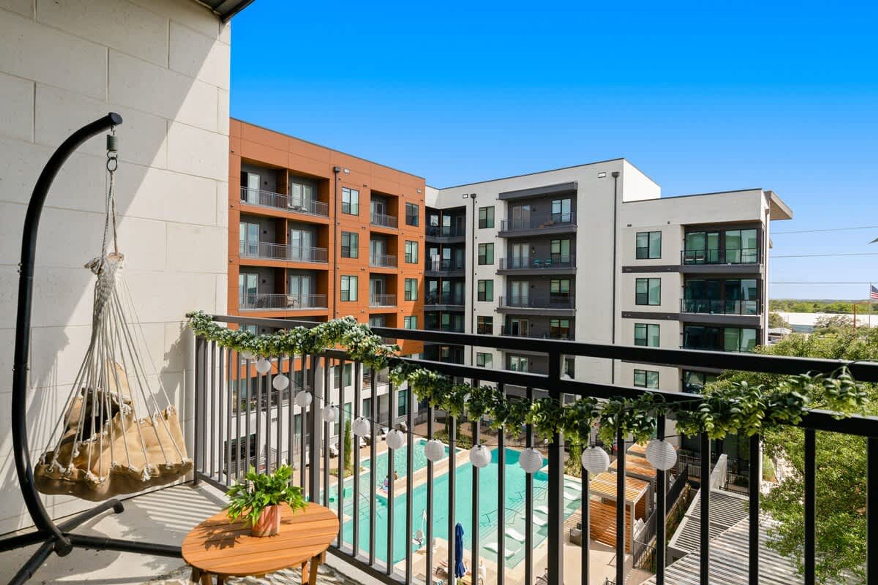 Un balcon avec un fauteuil suspendu, une petite table avec une plante, et une vue sur une piscine et des immeubles à plusieurs étages.