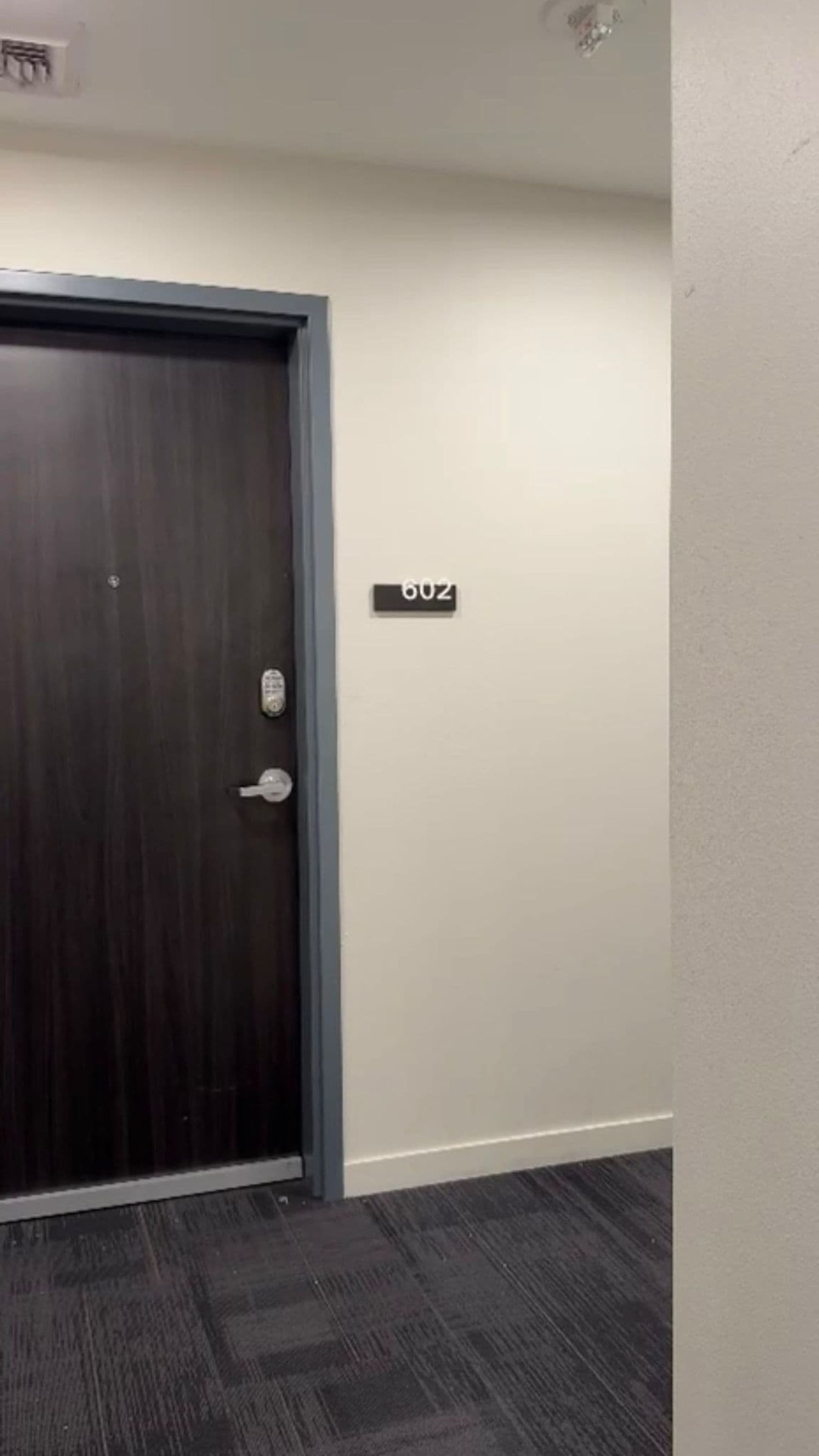 Una puerta de madera oscura con un ojo de cerradura y un cerrojo, un cartel con el número de la puerta, y un piso alfombrado.