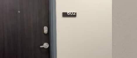Una puerta de madera oscura con un ojo de cerradura y un cerrojo, un cartel con el número de la puerta, y un piso alfombrado.