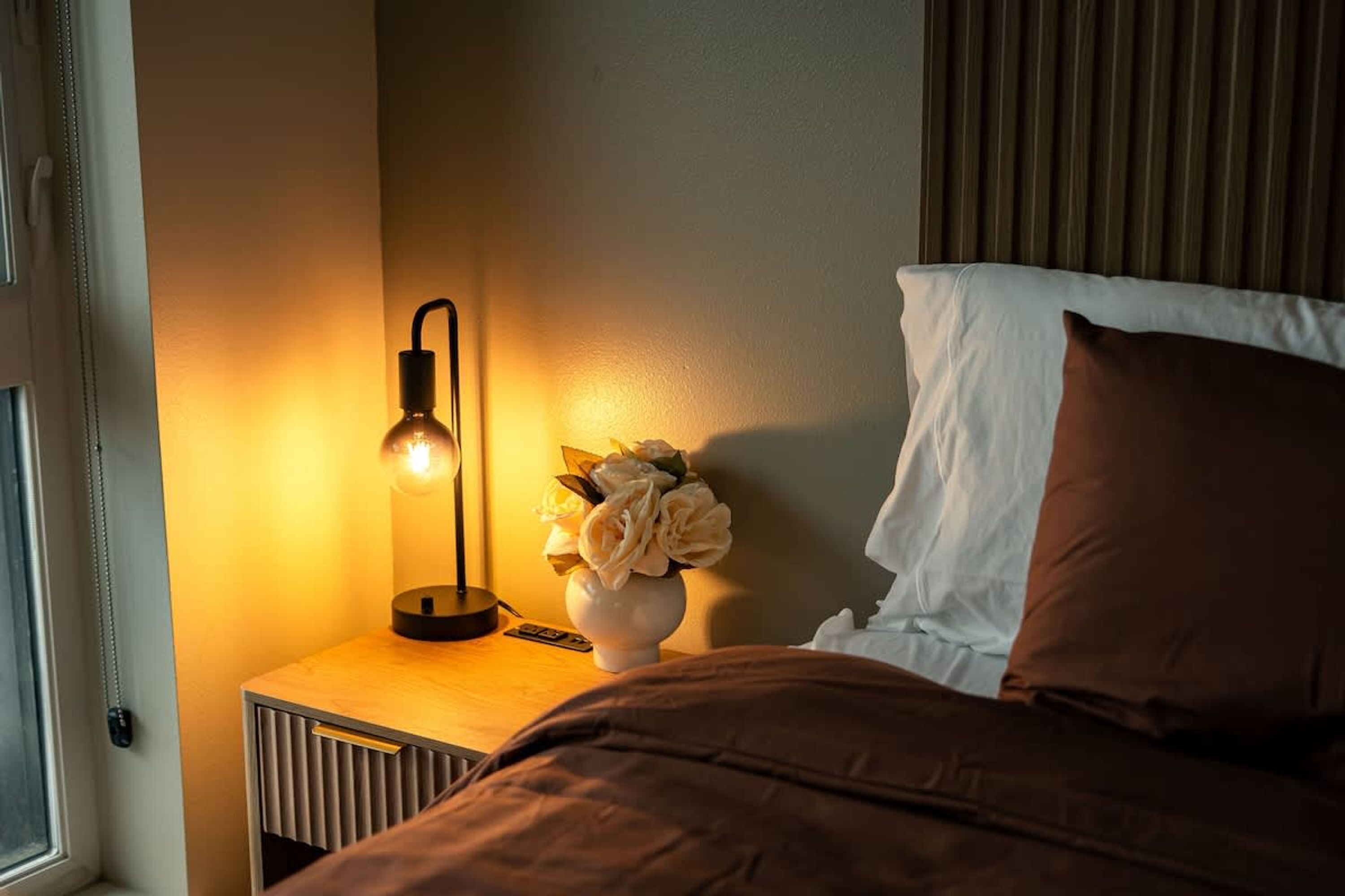 A bedroom with a bedside lamp, a vase with flowers, a bed with brown bedding, and a window with blinds.