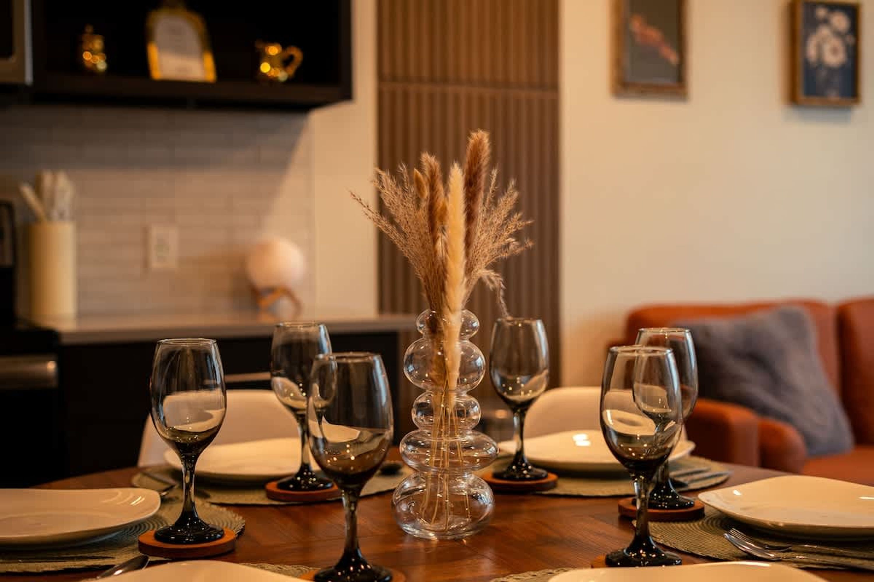 A dining table set with wine glasses and plates, a decorative vase with dried plants, and a modern kitchen in the background.