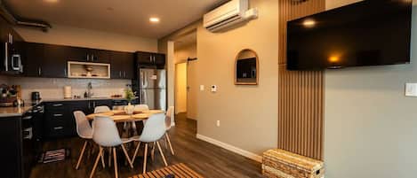 A modern kitchen with dark cabinets, a dining area with a wooden table and chairs, a flat-screen TV mounted on the wall, and a small wooden chest.