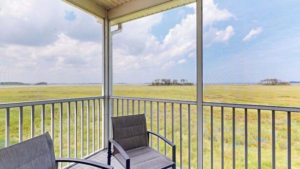 A covered balcony with two chairs, a view of a grassy field, and a clear sky. - Charming condo with nearby golf, outdoor pool & close to the beach (Ocean View)