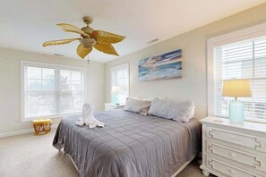 Ein Schlafzimmer mit einem Bett, Deckenventilator, einem Fenster mit Jalousien, einem Nachttisch mit Lampe und einem kleinen Hocker.
