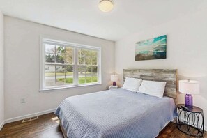 A bedroom with a large window, a bed with white bedding, a wooden headboard, and a wall-mounted painting. - Holy Park house w/deck, patio, grill, close to beach & restaurants. (Ocean View)
