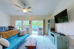 A living room with a blue sofa, a wooden coffee table, a ceiling fan, a television on the wall, and a sliding glass door leading to an outdoor area. - Townhomes of Providence spacious townhouse w/shared pool, deck, W/D (Ocean View)