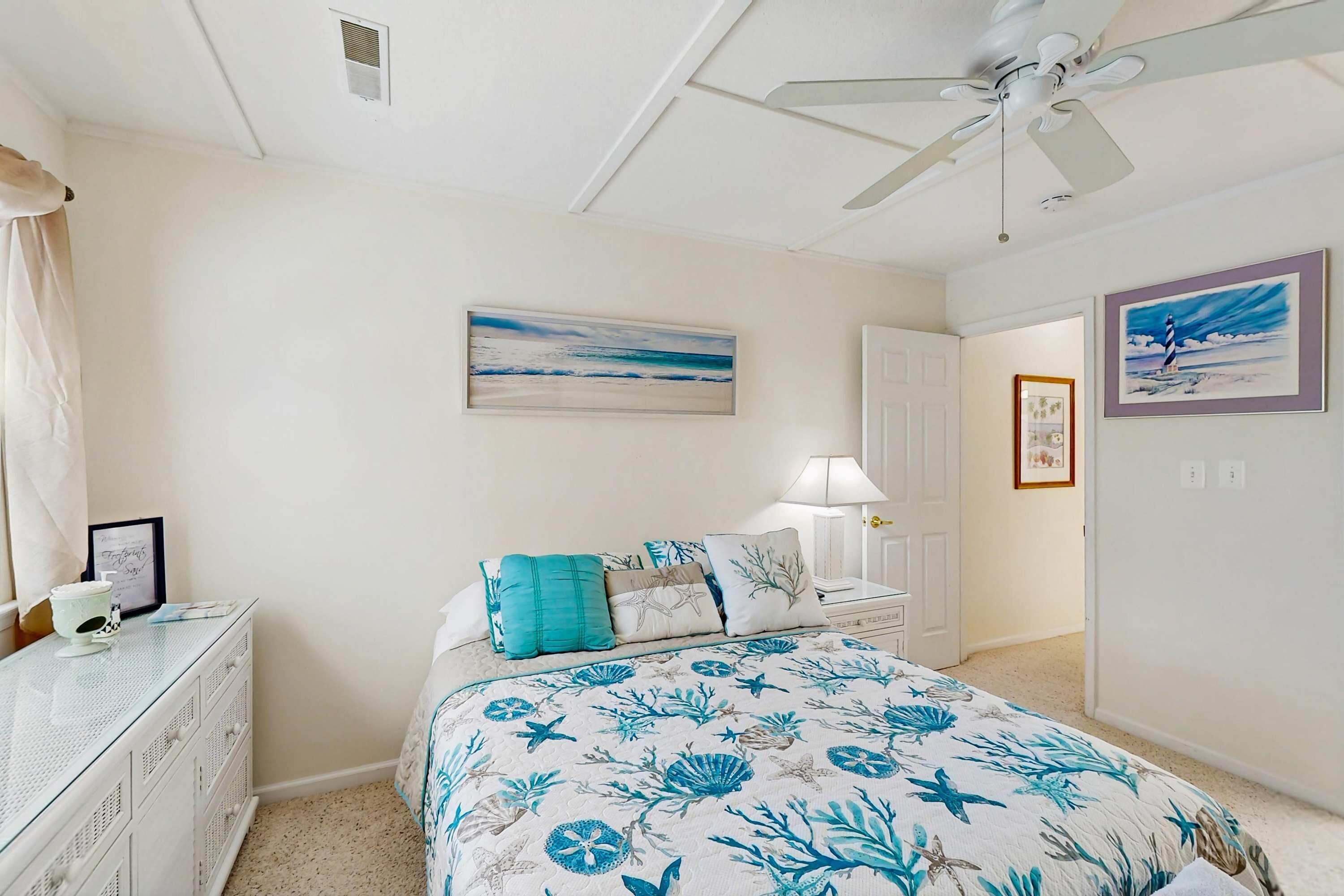 Une chambre à coucher comprenant un lit, une table de chevet, une commode, un ventilateur de plafond et une porte donnant sur une autre pièce.