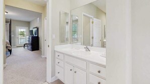 Une salle de bain avec un grand miroir, un meuble-lavabo blanc et une porte donnant sur une autre pièce.