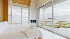 A bedroom with a large bed, a wooden ceiling, and a view of a water body through large windows. - Bayview Park bay front house w/private pool, amazing bay views, deck, balcony (Bethany Beach)
