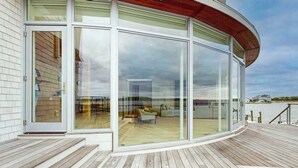 A modern building with large glass doors, a wooden deck, and a view of a beach. - Bayview Park bay front house w/private pool, amazing bay views, deck, balcony (Bethany Beach)
