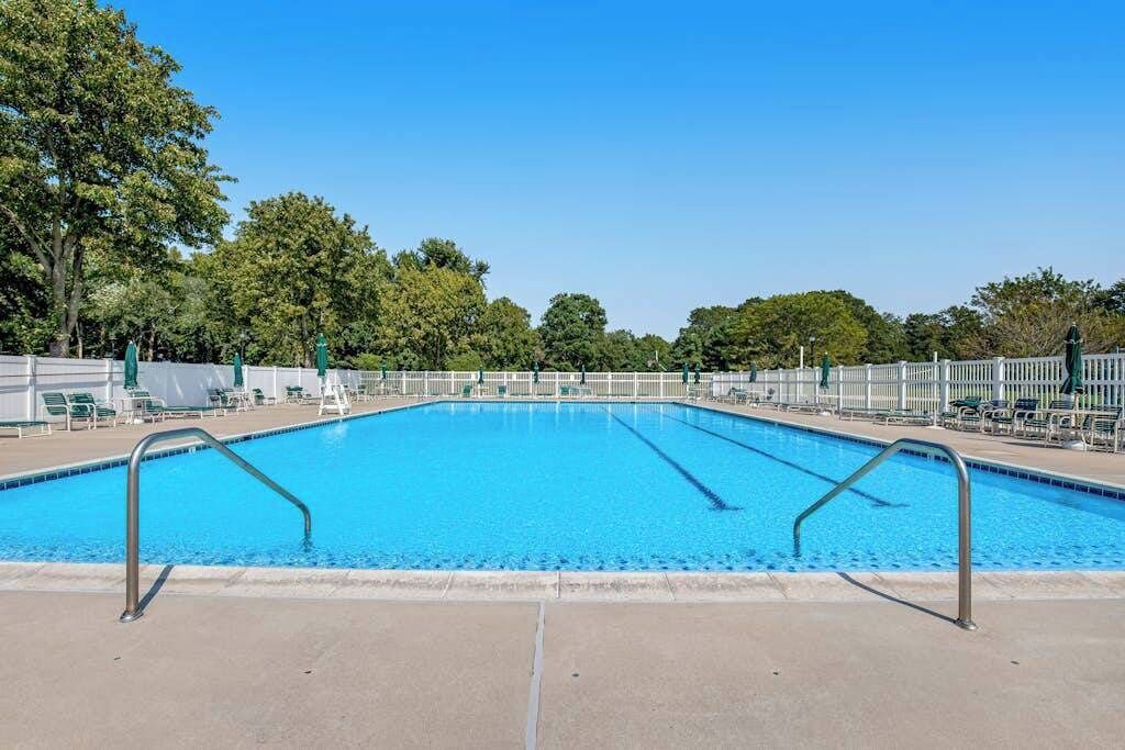 Una piscina rectangular con agua azul cristalina, rodeada por una cerca blanca y árboles al fondo.
