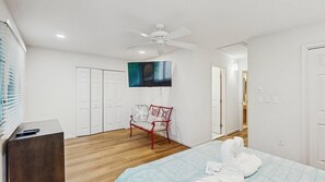 A bedroom with a bed, a chair, a TV, a ceiling fan, and a wooden floor.