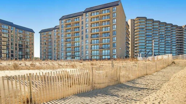 A high-rise residential building with multiple balconies, a sandy beach in the foreground, and a clear blue sky. - Sea Colony oceanfront 7th-floor condo with pool & tennis/basketball courts (Bethany Beach)