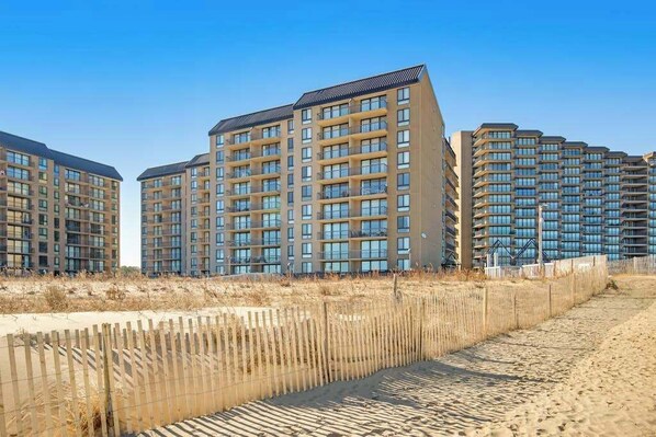 A high-rise residential building with multiple balconies, a sandy beach in the foreground, and a clear blue sky. - Sea Colony oceanfront 7th-floor condo with pool & tennis/basketball courts (Bethany Beach)