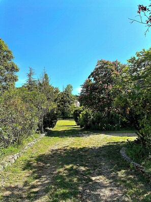 A pathway leading through a garden with various trees and shrubs, a clear sky, and a distant building. - 90m2 duplex apartment with 2000m2 enclosed garden (Ruoms)
