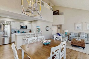 A spacious living area with a dining table, white chairs, a kitchen with modern appliances, and a loft bedroom with a balcony. - Sea Colony Tennis 2nd-floor condo w/community pools, tennis/pickleball, gym (Bethany Beach)