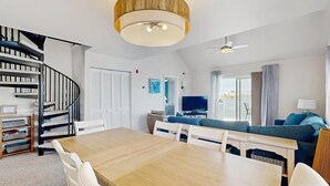 Ein modernes Wohnzimmer mit Esstisch aus Holz, weißen Stühlen, einem blauen Sofa, einem Fernseher und einer Wendeltreppe.
