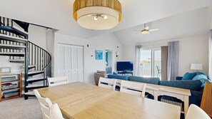Ein modernes Wohnzimmer mit Esstisch aus Holz, weißen Stühlen, einem blauen Sofa, einem Fernseher und einer Wendeltreppe.