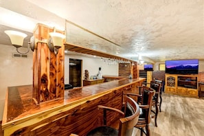 Angolo bar con bancone in legno e sgabelli, una televisione montata su un muro in mattoni e un mobile in legno con scaffali.