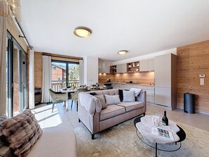 A modern living room with a sofa, a dining area with chairs, a kitchen with wooden cabinets, and a large window. - Rennes - Apartment - Sleeps 6 - Jacuzzi (Les Gets)