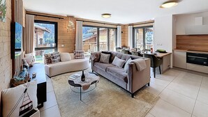 A spacious living area with a sofa, a coffee table, and a view of a mountain cabin through large windows. - Rennes - Apartment - Sleeps 6 - Jacuzzi (Les Gets)