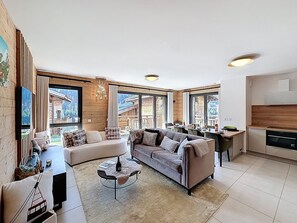 A spacious living area with a sofa, a coffee table, and a view of a mountain cabin through large windows. - Rennes - Apartment - Sleeps 6 - Jacuzzi (Les Gets)