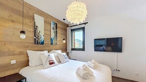 A bedroom with a wooden wall, a bed with white bedding, decorative pillows, a large fluffy light fixture, a TV mounted on the wall, and a window with a view. - Rennes - Apartment - Sleeps 6 - Jacuzzi (Les Gets)