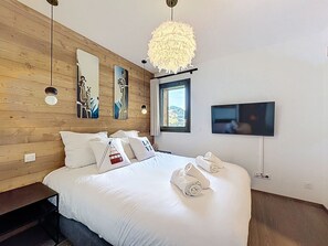 A bedroom with a wooden wall, a bed with white bedding, decorative pillows, a large fluffy light fixture, a TV mounted on the wall, and a window with a view. - Rennes - Apartment - Sleeps 6 - Jacuzzi (Les Gets)