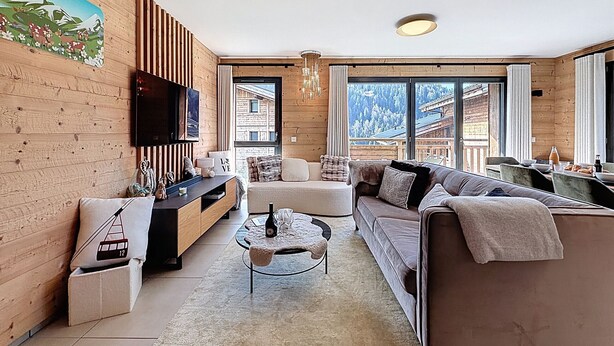 A modern living room with wooden walls, a flat-screen TV, a sofa, a coffee table, and a view of a mountainous landscape through large windows. - Rennes - Apartment - Sleeps 6 - Jacuzzi (Les Gets)