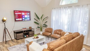 Ein modernes Wohnzimmer mit großem Flachbildfernseher, einer Couch, zwei Sesseln, einem Couchtisch und einer Zimmerpflanze.
