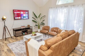 Ein modernes Wohnzimmer mit großem Flachbildfernseher, einer Couch, zwei Sesseln, einem Couchtisch und einer Zimmerpflanze.