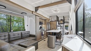 A modern RV interior with a sofa, kitchenette, dining area, and large windows. - The Pine- Smoky Mountains View Tiny House w/ Hot Tub & Deck (Tallassee)