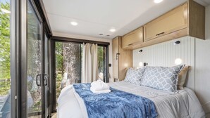 Ein Schlafzimmer mit einem Bett, einer blauen Decke, einem weißen Handtuch, einer Holzkommode und Blick ins Grüne.