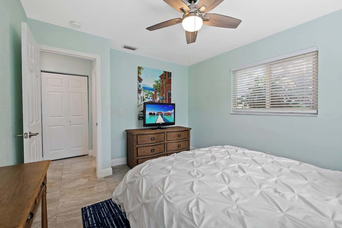 A bedroom with a bed, a television, a ceiling fan, a door, and a window with blinds.