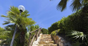 Eine steinerne Treppe mit Metallgeländer, umgeben von üppiger Vegetation und blauem Himmel.