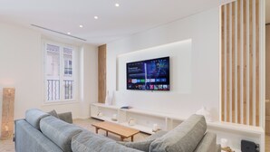 Una sala de estar moderna con un sofá gris, una mesa de centro de madera y un televisor montado que muestra la interfaz de un servicio de streaming.