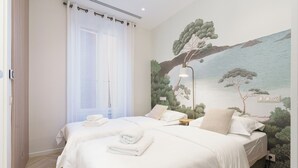 Habitación de hotel con dos camas, un mural grande de paisaje costero, una ventana con cortinas traslúcidas y una mesita de noche con lámpara.