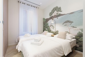 Ein Hotelzimmer mit zwei Betten, einer großen Küstenlandschaft als Wandbild, einem Fenster mit durchscheinenden Vorhängen und einem Nachttisch mit Lampe.
