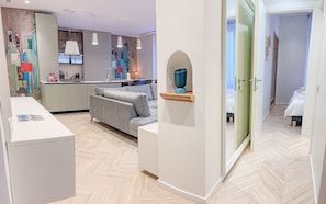 Ein modernes Hotelzimmer mit Sofa, einem kleinen Tisch, einer Küchenzeile und einem durch eine offene Tür einsehbaren Badezimmer.