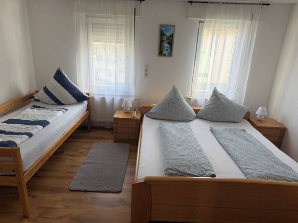 Una habitación con dos camas de madera, una mesita de noche con lámpara, un tapete gris y una ventana con cortinas translúcidas.