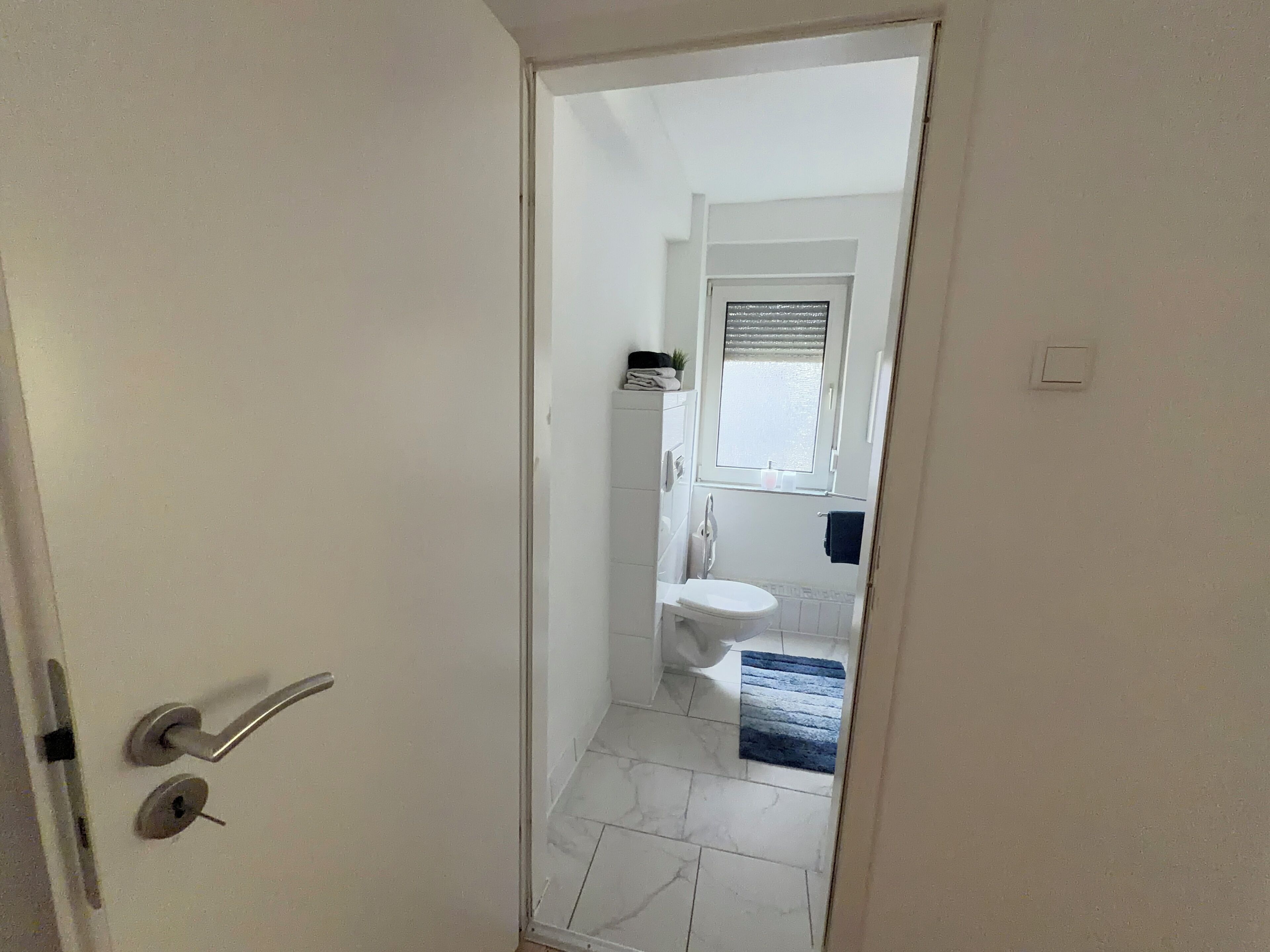 A narrow hallway leading to a bathroom with a toilet, a small window, and a towel rack.