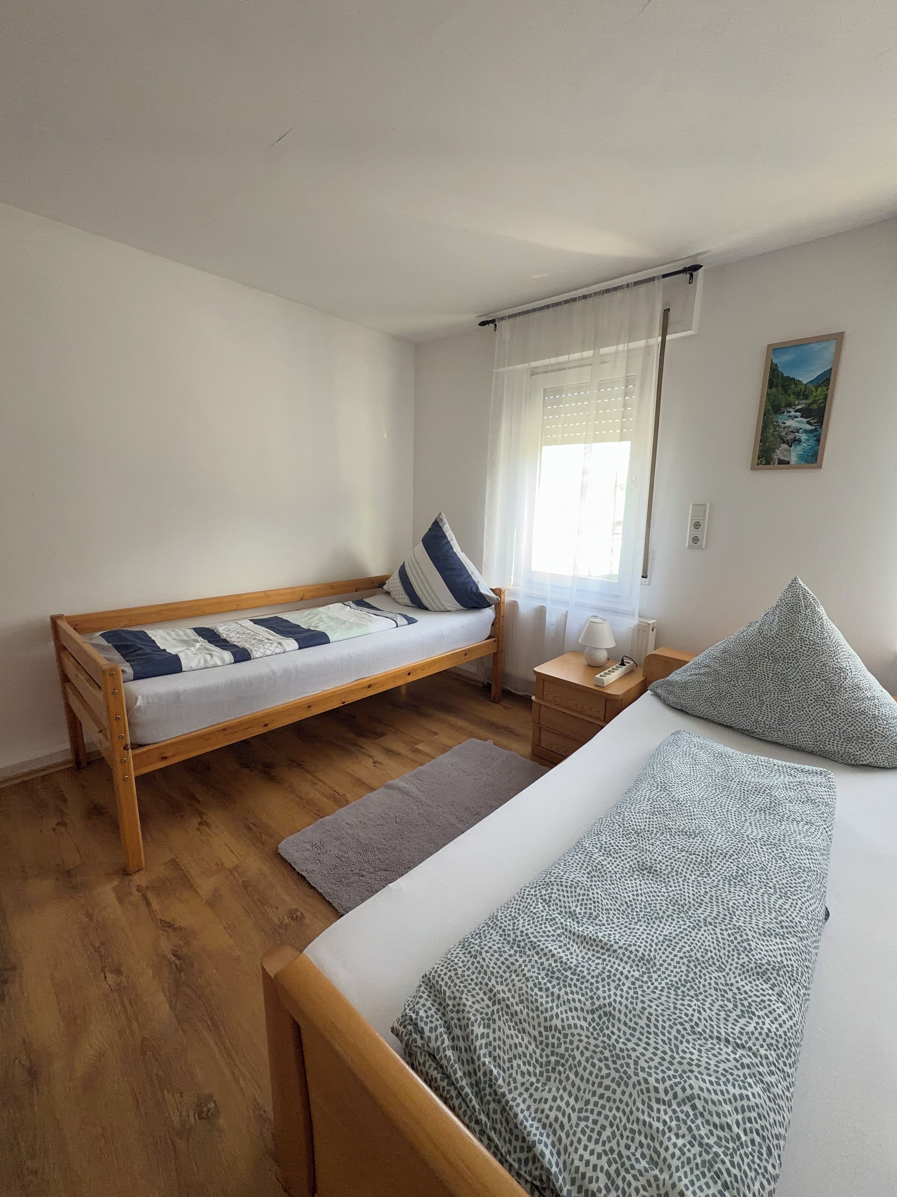Una stanza con un letto in legno, un letto più piccolo, un comodino, una lampada, un quadro al muro e una finestra con tende.