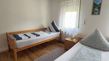 Una stanza con un letto in legno, un letto più piccolo, un comodino, una lampada, un quadro al muro e una finestra con tende.