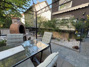 Une terrasse avec une table en verre, deux chaises, un four en briques et un jardin agrémenté de plantes et d’un mur.