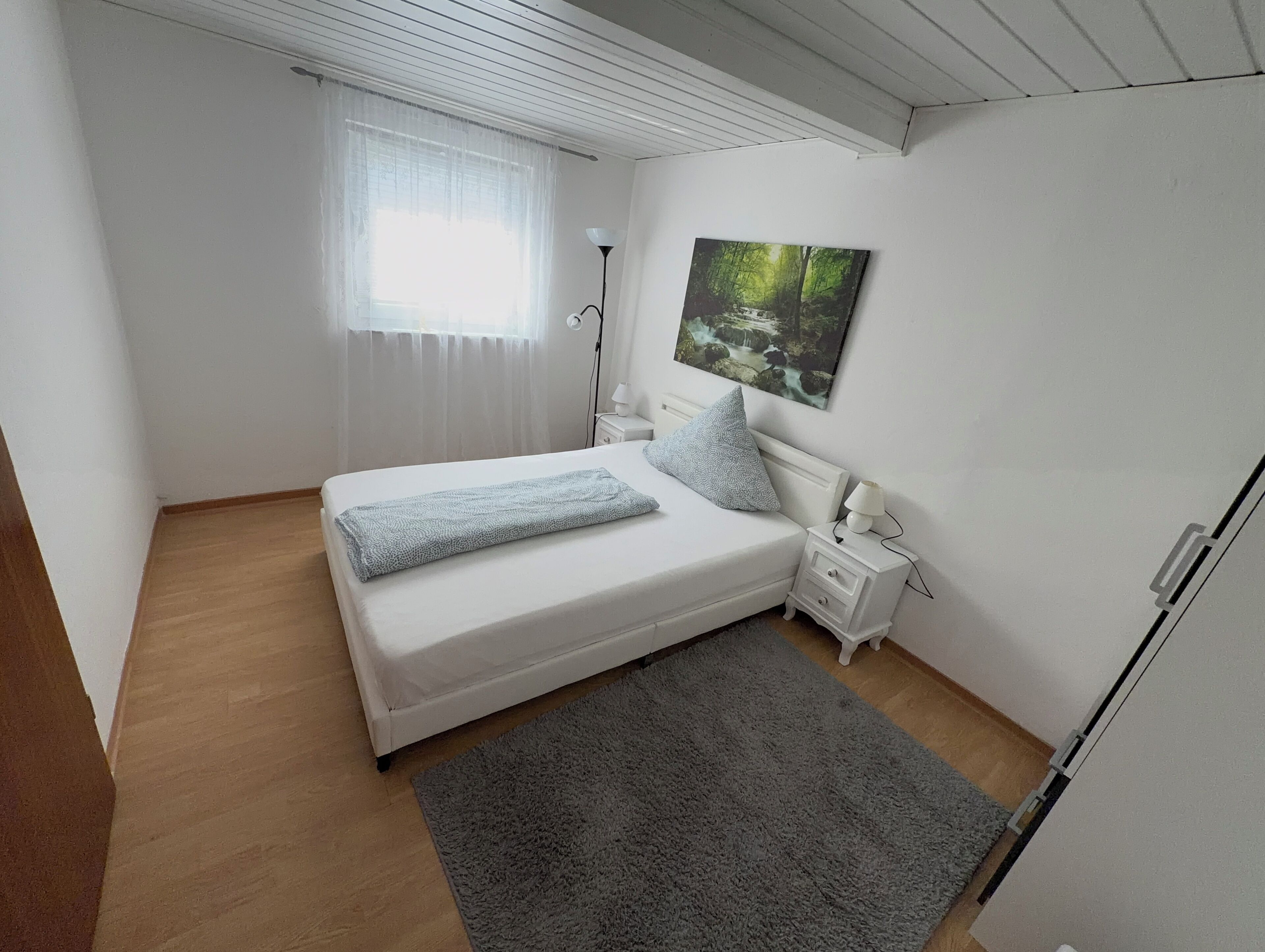 Ein Schlafzimmer mit Bett, Nachttischen, einer Lampe, einem Gemälde und einem Fenster mit Vorhängen.