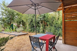 En terrasse med et bord og stole under en stor parasol, omgivet af en have med forskellige planter og et træhegn.