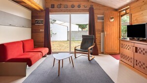 Ein Zimmer mit einem roten Sofa, einem Holzstuhl, einem kleinen weißen Tisch, einem Fernseher auf einer Holzkommode und einer Glastür, die nach draußen führt.
